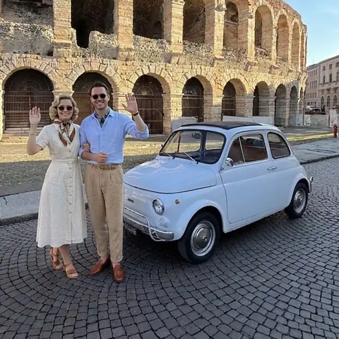 Una coppia in abito d'epoca saluta accanto alla Fiat 500 bianca davanti agli archi dell'Arena